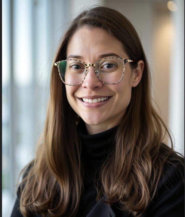 A person with brown hair and glasses smiles indoors in a well-lit, modern environment. The background shows blurred windows.