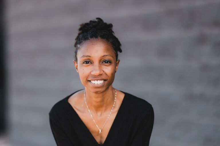 A person with a black shirt smiles against a blurred gray brick wall background. No recognizable landmarks or historical buildings are visible.