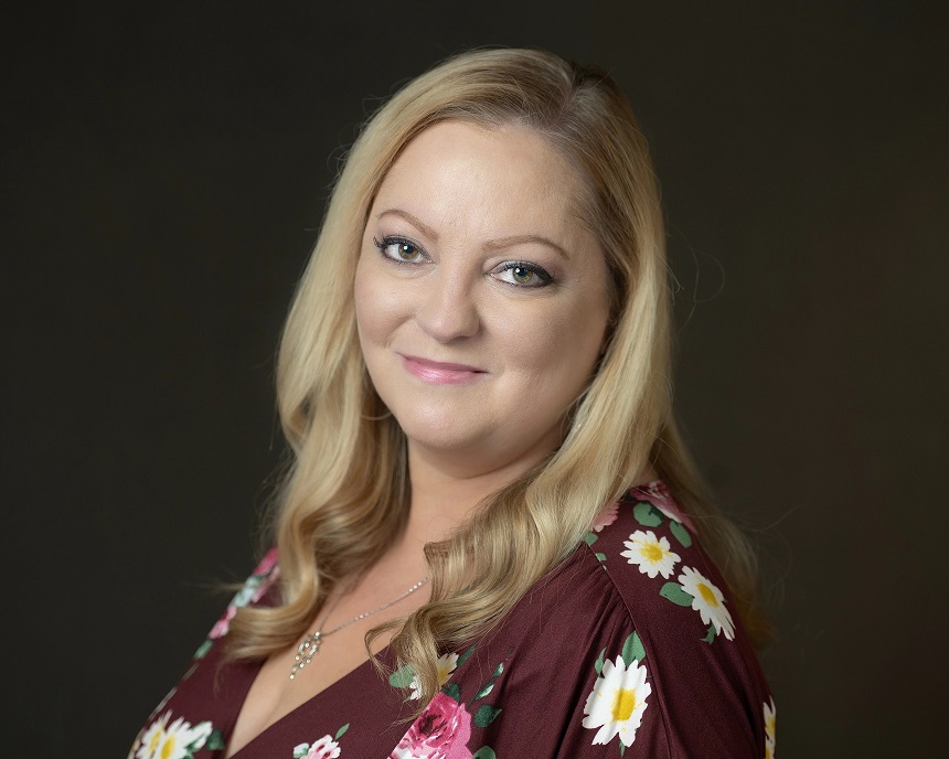 A person with long blonde hair smiles, wearing a floral dress against a dark background. Close-up portrait with a calm, friendly expression.