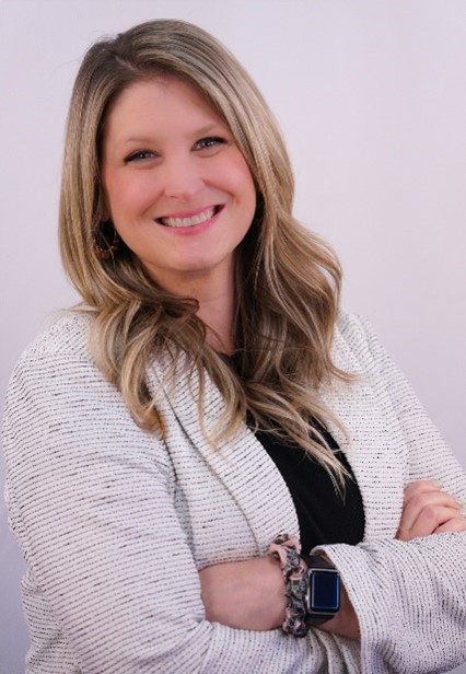 A smiling person with long hair and a light sweater stands against a plain background, with arms crossed, wearing a smartwatch and bracelet.