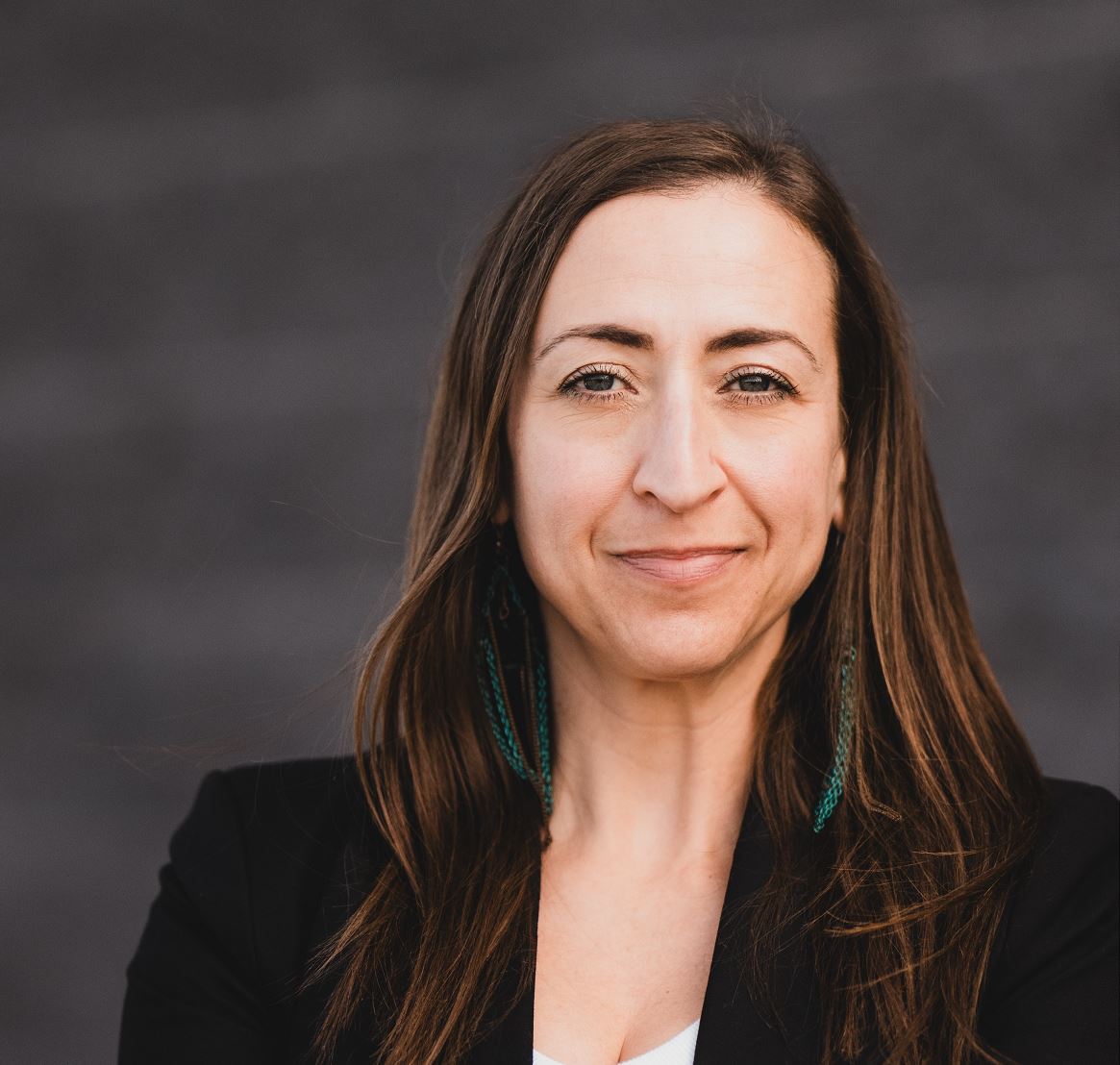 A person with long hair and earrings smiles at the camera, set against a plain, dark background.