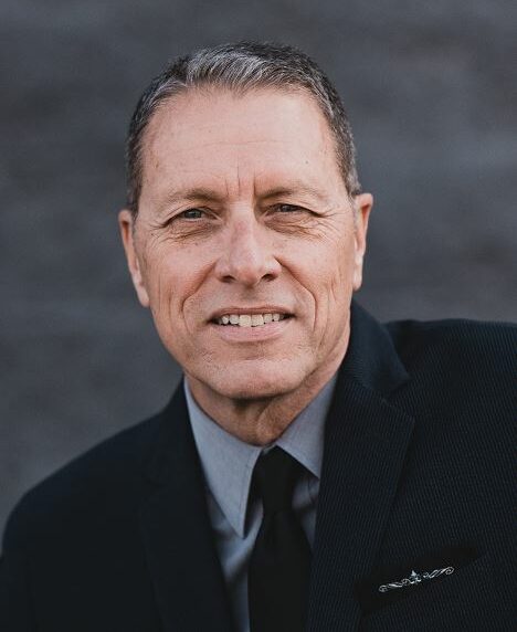 A person in a suit poses against a plain, gray background, smiling slightly, showcasing a professional portrait style with high contrast lighting.