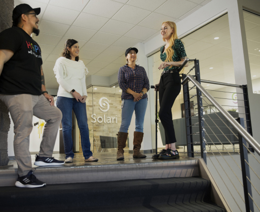 Four people stand on stairs, engaging in conversation. The setting is modern and indoor, with a sign reading "Solari" visible in the background.