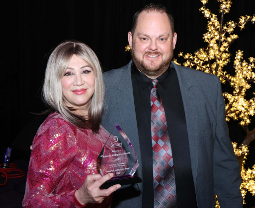 Two persons posing at an event. One holds an award. They stand in front of a decorative, illuminated tree on a dark background.