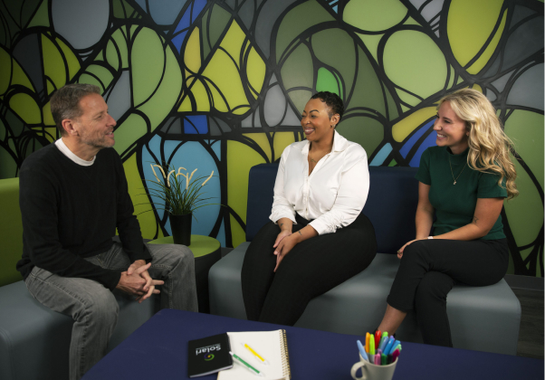 Three people are sitting on blue chairs, engaged in conversation. Colorful abstract mural and office supplies, including notebooks and pens, are visible.