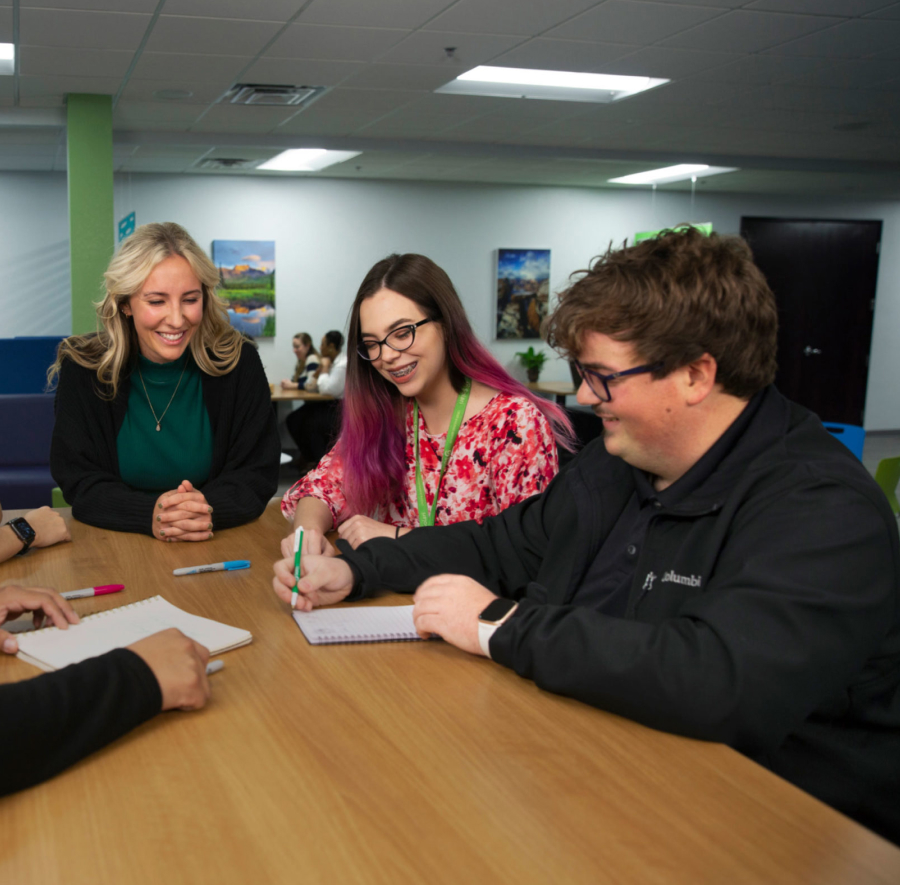 Four people collaborate at a table in an office setting, surrounded by framed landscape photos. They appear engaged and focused.