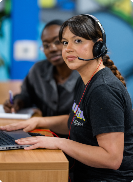 Two people working together, one wearing a headset, using a laptop. They are in a casual, indoor setting with colorful decor in the background.