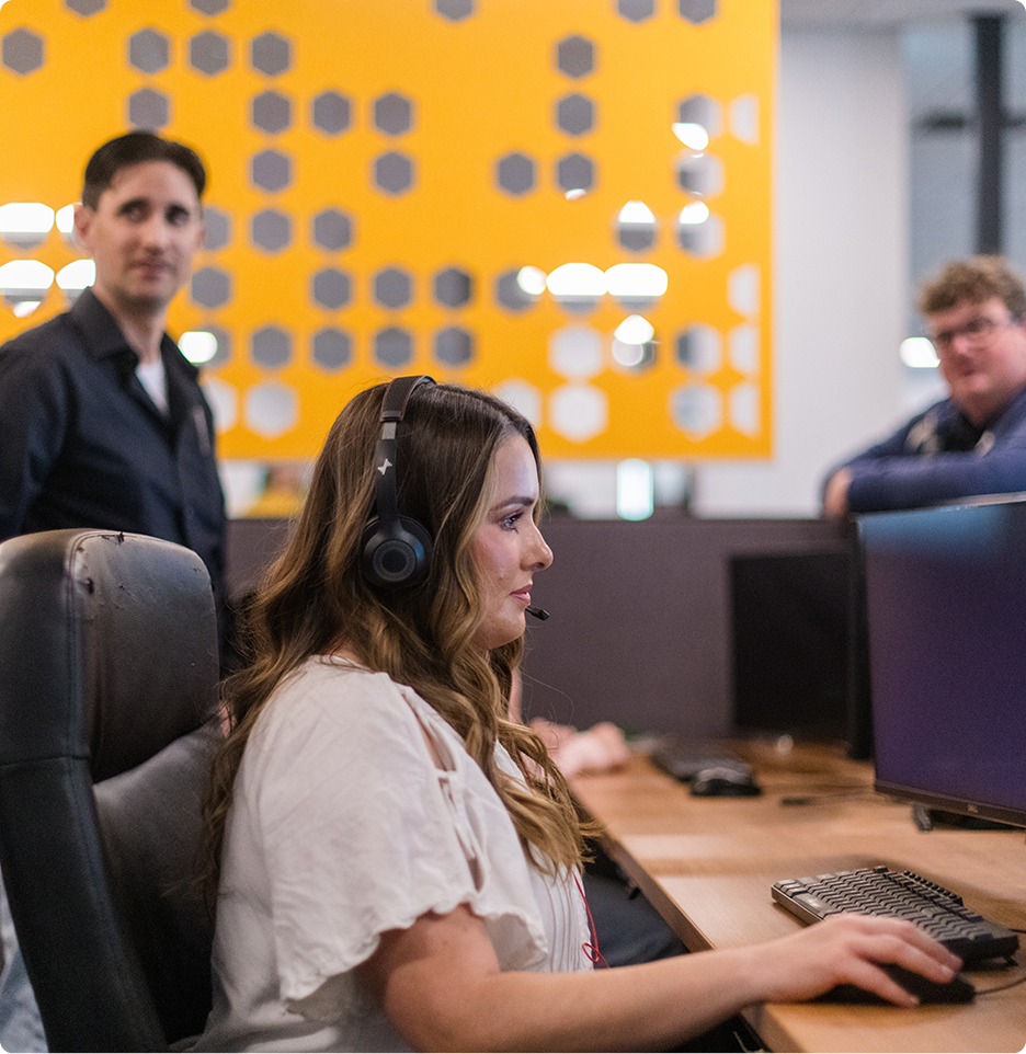 A person wearing headphones works at a computer in an office, while two other people look on, against a geometrically decorated wall.