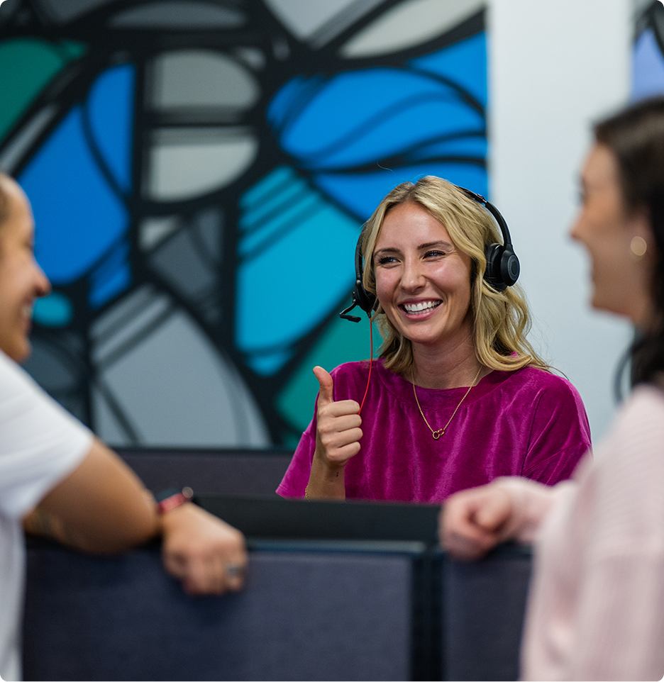 A person wearing a headset smiles and gives a thumbs-up, engaging with others in a modern, colorful office setting.