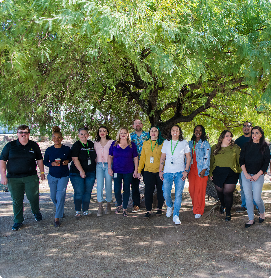 A group of eleven people stands together outdoors under a large tree, smiling, with a natural, sunny setting in the background.