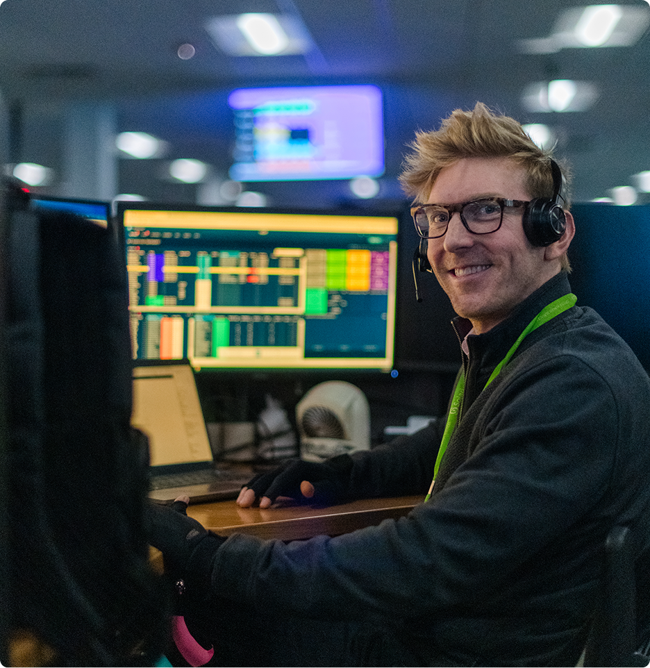 A person with a headset sits at a computer station, displaying complex data on the screens, smiling in a tech-focused office environment.