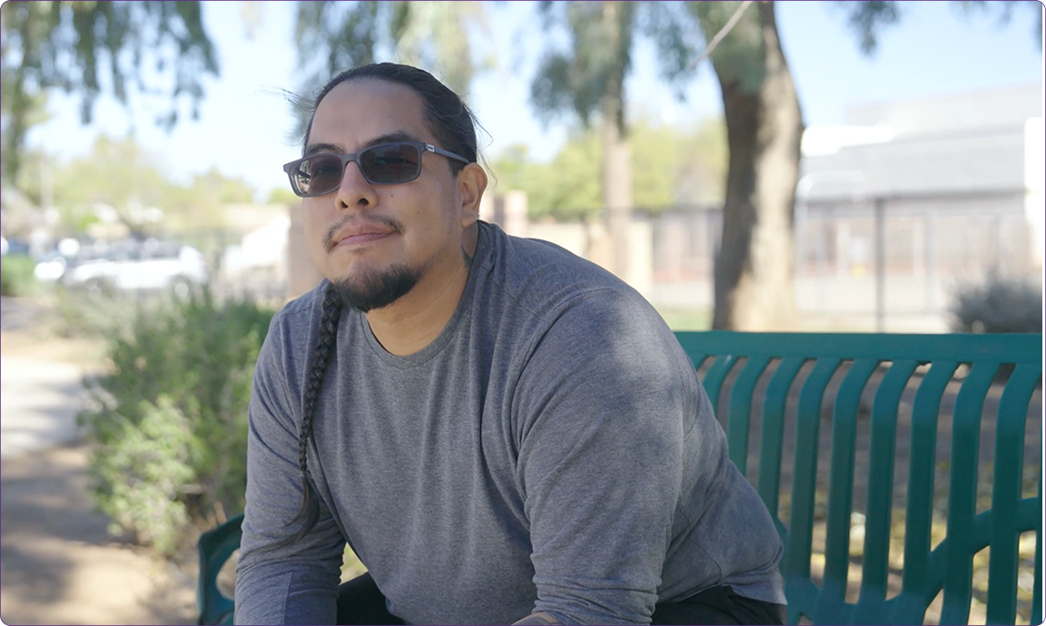 A person wearing sunglasses and a gray shirt sits on a green bench in a park, surrounded by trees and blurred background.