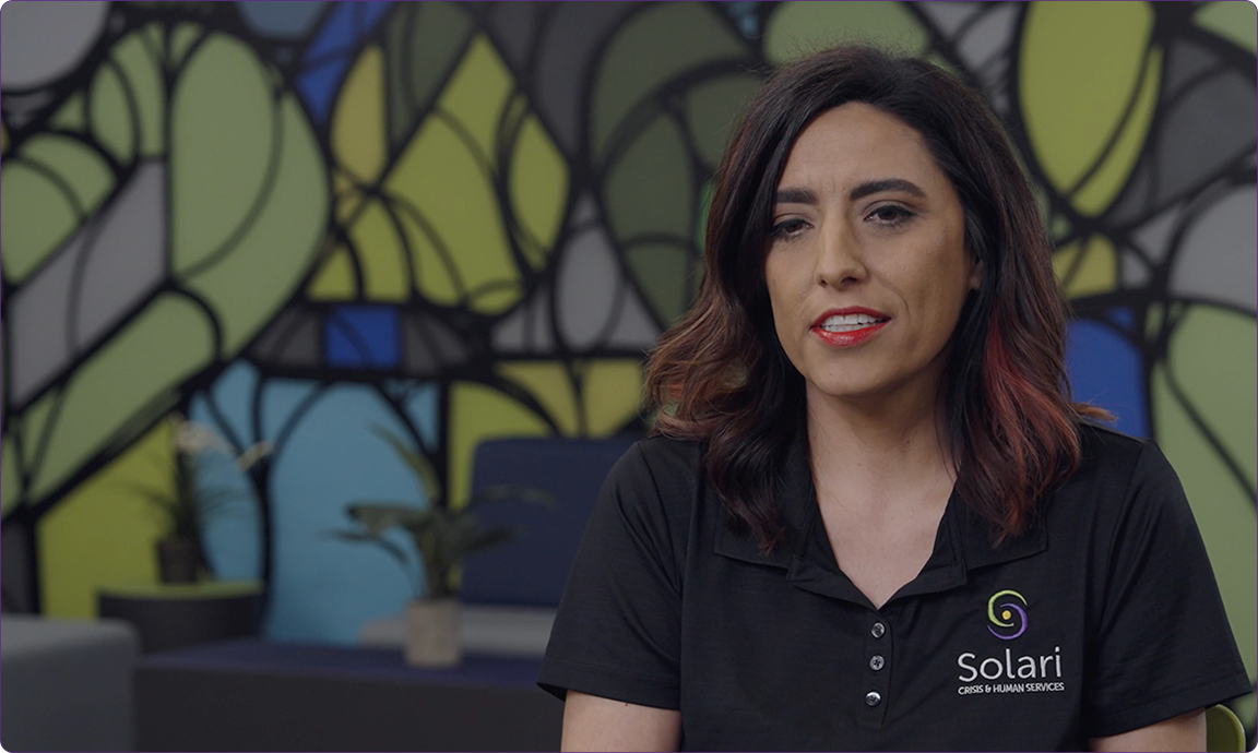 A person wearing a black "Solari Crisis & Human Services" shirt sits in front of a colorful, geometric wall design, with plants visible.