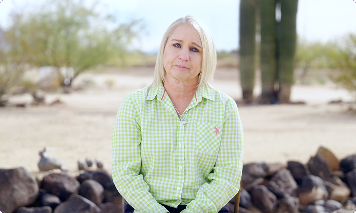 A person in a green checkered shirt sits outdoors, with rocks and desert vegetation, including cacti, in the background.