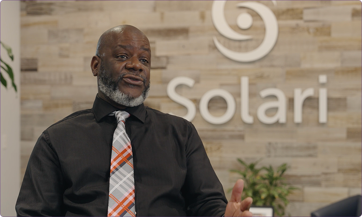 A person wearing a patterned tie sits indoors, gesturing while speaking. The word "Solari" is displayed on the wall behind them.