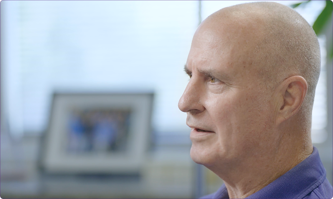 A person with a shaved head, wearing a blue shirt, is in profile view in a room with a blurred framed photo.