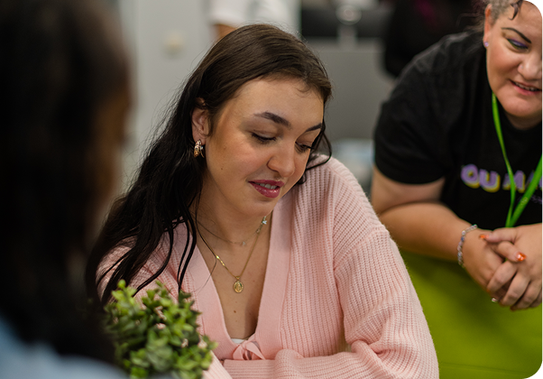 Three people gathered indoors, one looking at a small plant on the table. Casual, relaxed setting with friendly interaction.