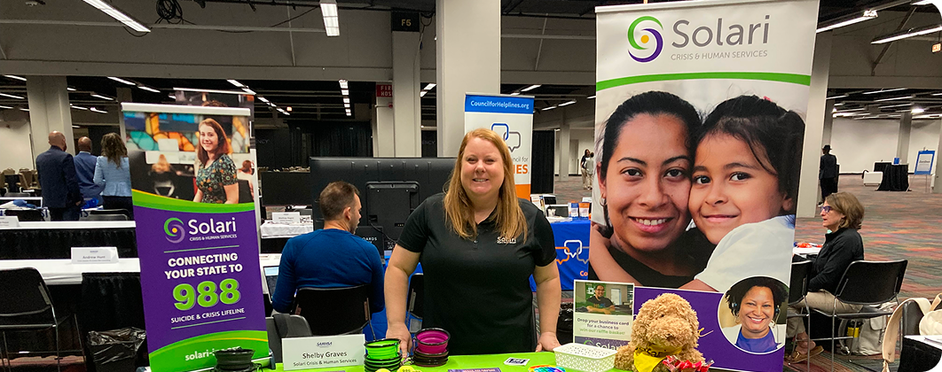 A person stands behind a Solari Crisis & Human Services booth at an indoor event, surrounded by informational banners and promotional materials.