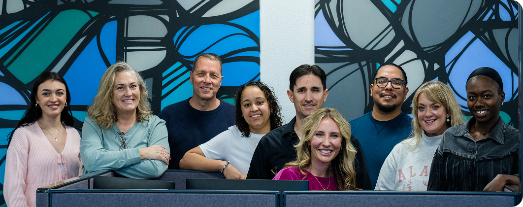 A group of nine people smiling together in an indoor setting, standing before a colorful, abstract mural featuring blue and black patterns.