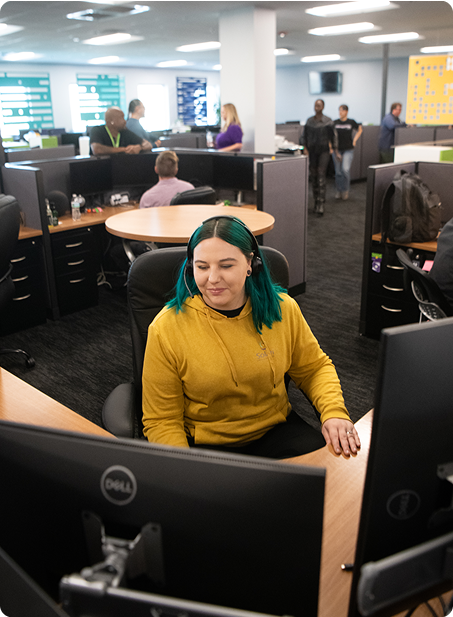 A person with headphones sits at a computer in a modern office with several other people working in the background.