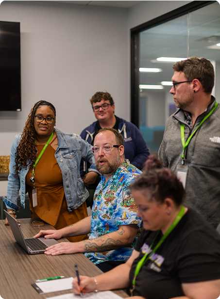 Five people gathered around a table in a modern office, engaged in discussion, with a laptop and notes visible on the table.