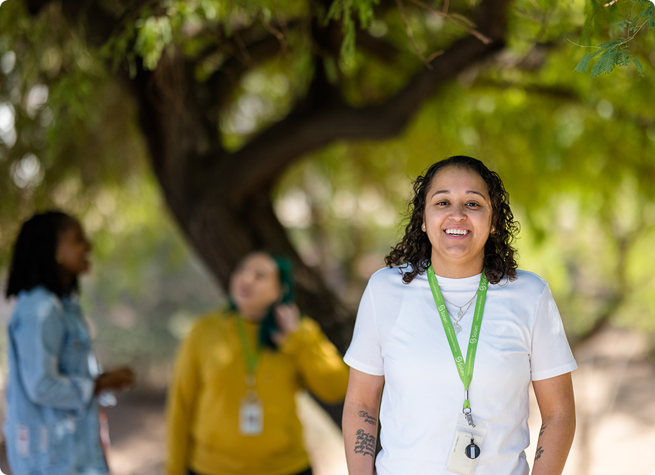 A person smiling in a park setting, wearing a lanyard, with two blurred individuals in the background near a tree.