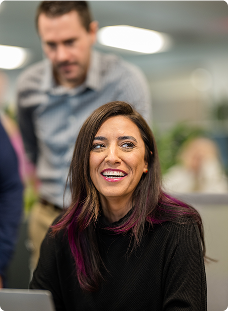 Two people in an office setting smiling and interacting, with blurred background suggesting teamwork or collaboration in a professional environment.