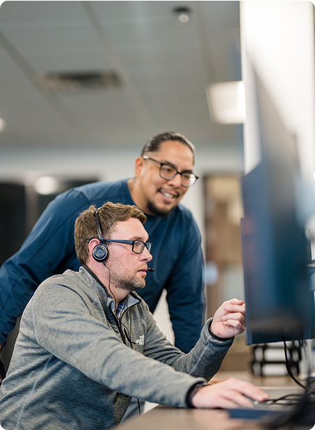 Two people in an office setting, one seated with headphones using a computer, the other standing, engaged and observing the screen.