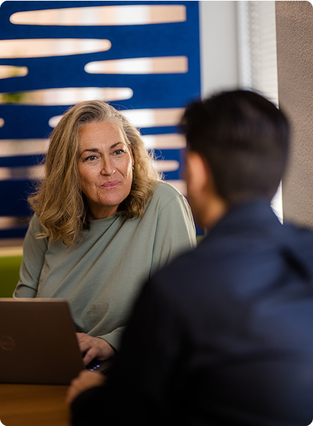 Two people are conversing indoors at a table. One person is using a laptop. There's a decorative blue partition in the background.