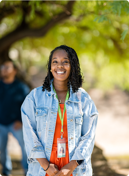 A person in a denim jacket smiles outdoors, with trees in the background. Another person is blurred behind them.