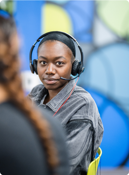 A person wearing a headset sits indoors, facing the camera. The background features colorful, blurred shapes, suggesting a modern, creative environment.