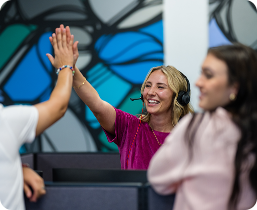 A person with a headset gives a high-five to another. Another person stands nearby, with a colorful abstract mural in the background.