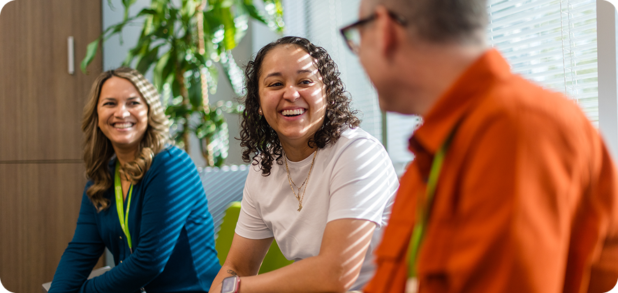 Three people sitting indoors, engaged in a lively conversation, smiling. Sunlight filters through blinds, with plants and office furniture in the background.