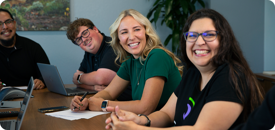 Four persons are gathered around a table, smiling and engaged. Papers and laptops are present, indicating a friendly meeting or discussion.