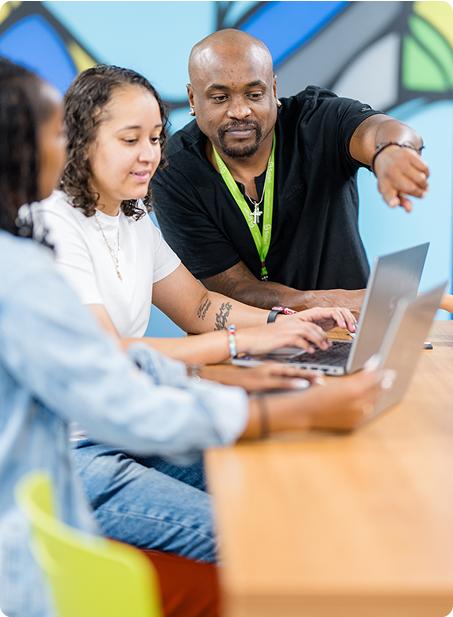 Three people collaborate at a wooden table, using laptops. One person points, guiding the others. Bright, abstract wall art in the background.