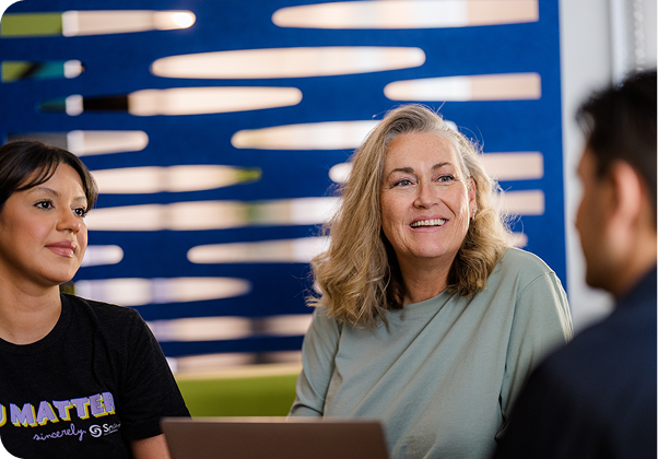 Three people are conversing indoors, seated with a laptop. The background features a modern, artistic blue panel design.