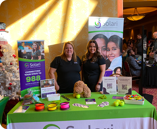 Two people stand behind an information table for Solari Crisis & Human Services, featuring promotional items and banners, at an indoor event.