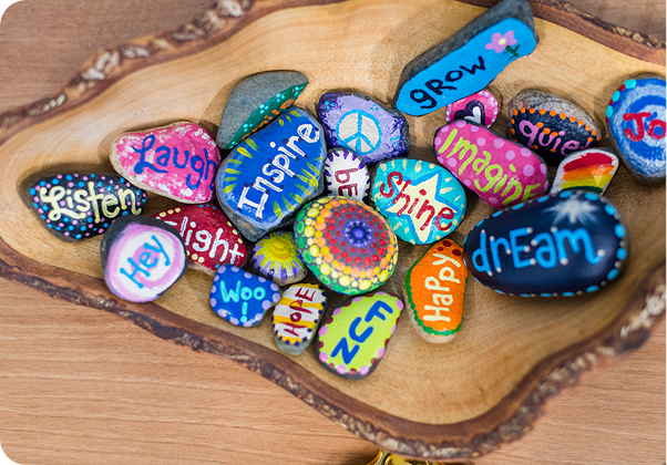 A wooden bowl holds colorful painted rocks with words like "Inspire" and "Dream," creatively decorated with vibrant patterns and symbols.