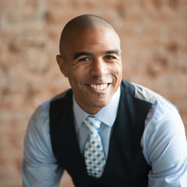 A person in formal attire smiling in front of a blurred brick wall background. The person wears a checked tie and dark vest.