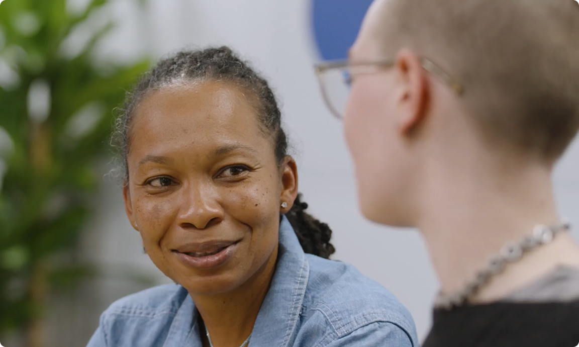 Two people are engaged in a conversation, smiling. Indoor setting with a green plant in the background, conveying a friendly atmosphere.