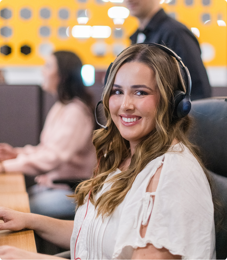 A smiling person with headphones sits at a desk, while others work in a modern office setting with bright decor.