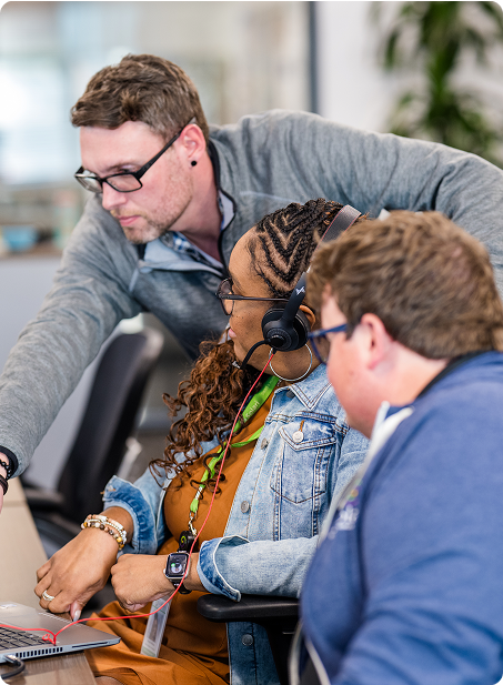 Three people collaborating at a desk, intently focused on a laptop. One person points at the screen, engaging in discussion or problem-solving.