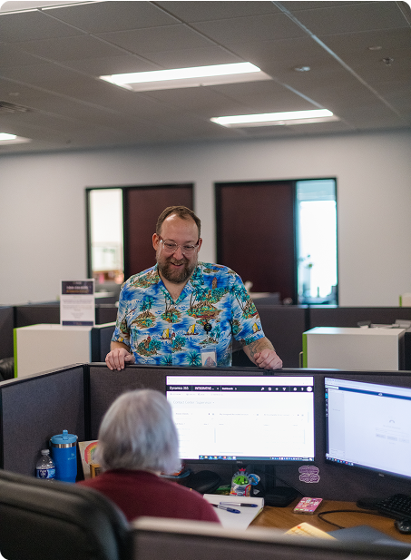 Two people in an office setting, interacting at a cubicle. One stands, wearing a colorful shirt, while the other sits at a computer.