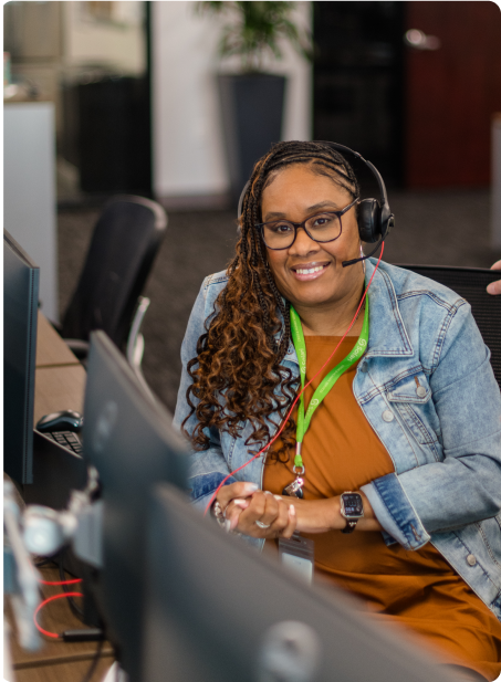 A person wearing a headset smiles while sitting at a desk in an office, interacting with a computer. Office environment is visible.